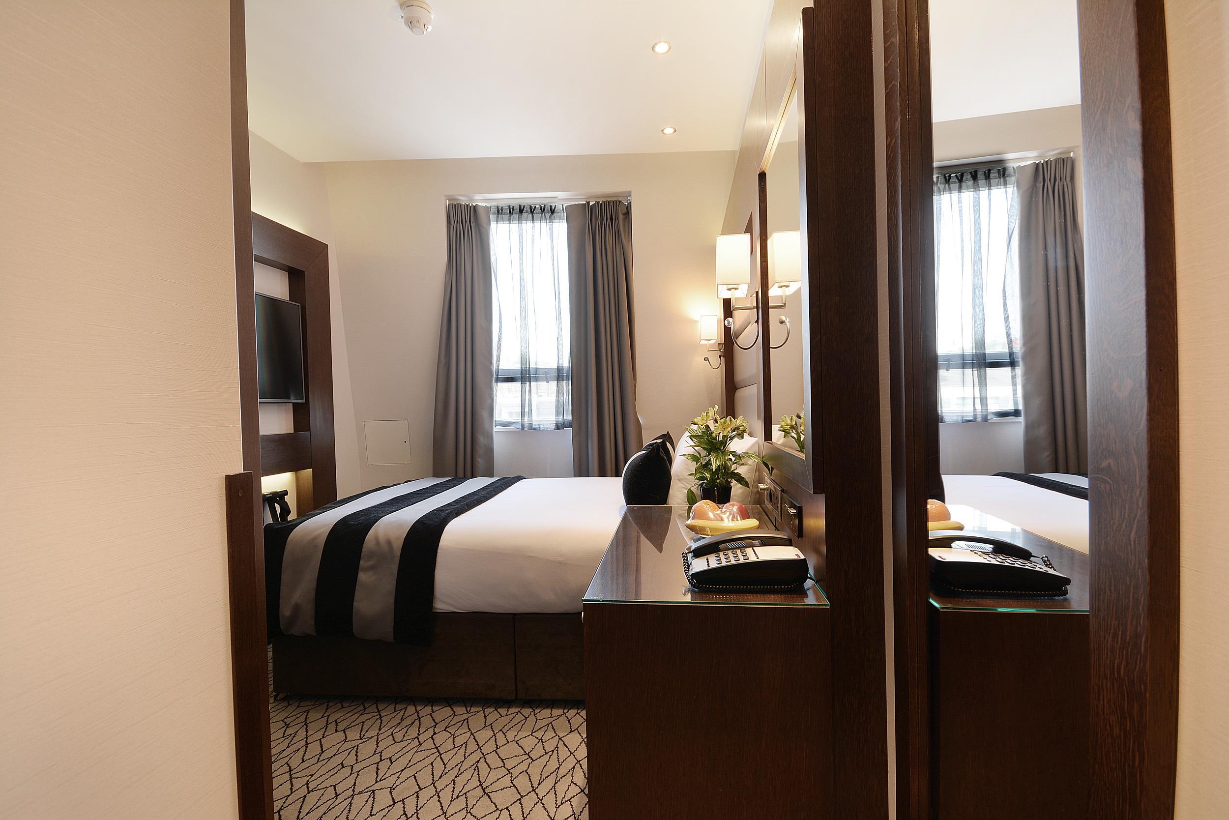Inhabit Queen'S Gardens, A Member Of Design Hotels London Eksteriør billede The photo shows a well-appointed hotel room. It features a neatly made bed with dark-striped bedding and a headboard, positioned against a wall. On either side of the bed, there are table lamps providing soft lighting. The room has large windows dres