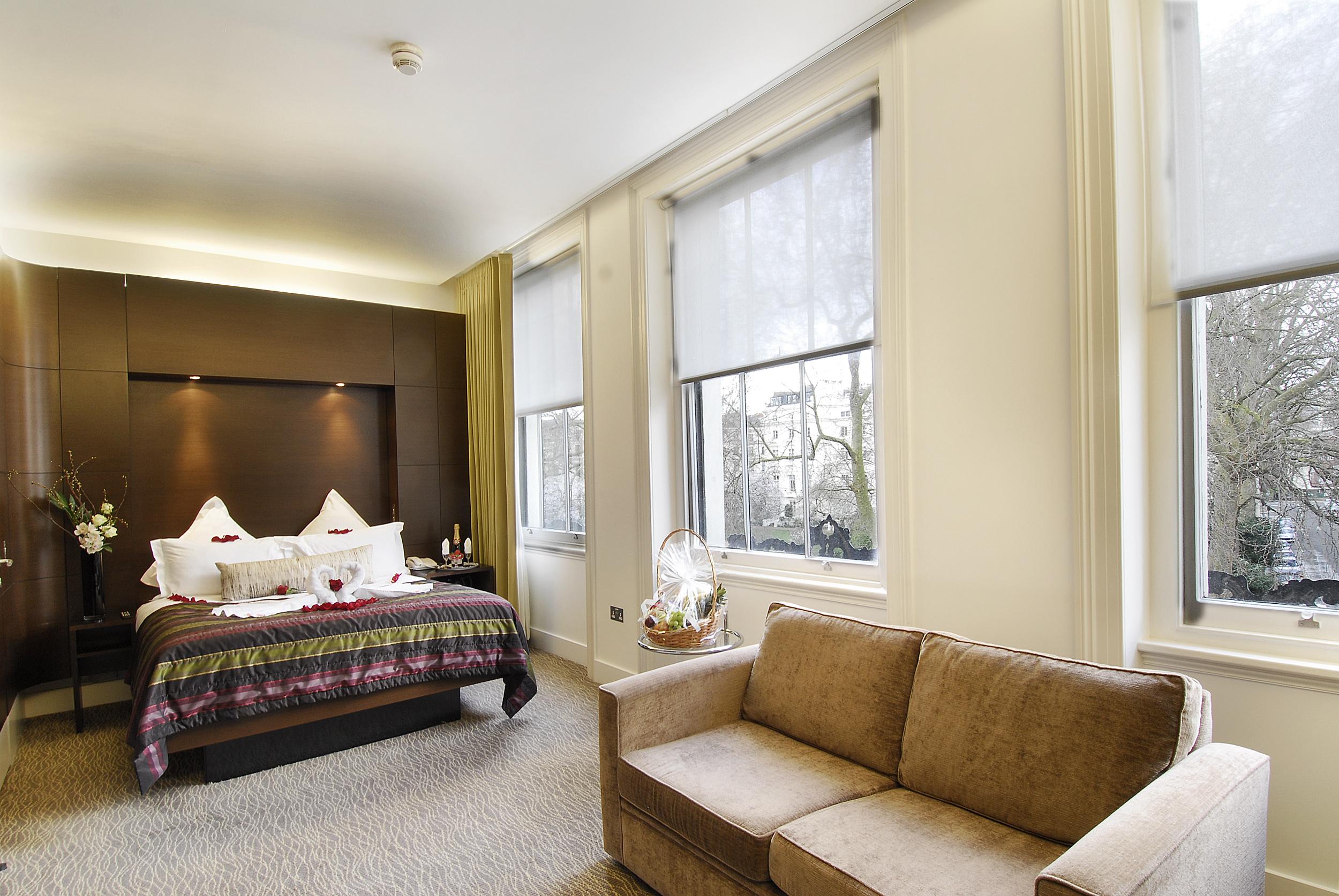 Inhabit Queen'S Gardens, A Member Of Design Hotels London Eksteriør billede The photo shows a cozy bedroom with a modern design. The room features a large bed adorned with decorative pillows and a colorful throw. Behind the bed is a dark accent wall with soft lighting. To the side, there's a comfortable beige sofa placed aga